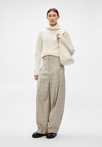 Camisola de gola alta creme em malha, calças de cintura alta aos quadrados bege e sapatos pretos de pele, segurando uma mala bege clara com ferragens minimalistas.