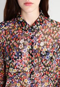 Transparent blommigt knapp-up skjorta i svart, med ett färgglatt mönster av blommor, en krage och två bröstfickor med knappstängningar.