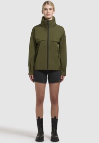 Veste vert olive avec col montant, fermeture éclair frontale et deux poches zippées. Conçue avec un tissu lisse etrésistant à l'eau.