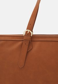 Bolsa tote sintética marrón con una textura suave, una correa plana con un hebilla de metal dorado y costuras precisas a lo largo de los bordes.