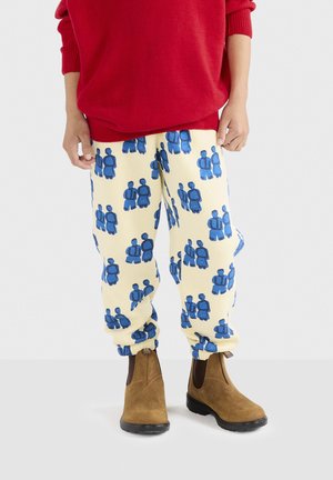 Pantalones de chándal de felpa en color crema con patrones de figuras humanas azules, con puños elásticos. Combinados con botas de tobillo marrones y un suéter rojo.