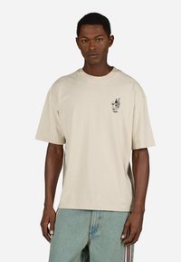 Bežs kokvilnas t-krekls ar īsām piedurknēm, apaļu kakla izgriezumu un mazu melnu grafiku kreisajā krūtī. Modelis valkā gaiši zilos džinsa bikses.