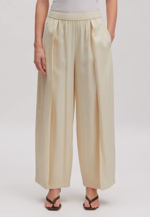 Frau trägt cremefarbene, plissierte Hosen mit weitem Bein und elastischem Bund sowie schwarze Zehensandalen, steht vor einem einfarbigen Hintergrund.
