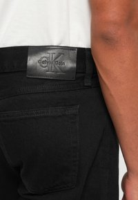 Crne traperice od denima s pravokutnom kožnom etiketom Calvin Klein straga, s klasičnim šavovima na džepovima i glatkom teksturom.