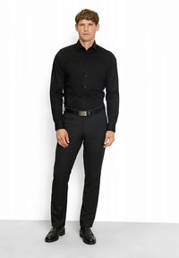 Camicia nera con bottoni e maniche lunghe, abbinata a pantaloni neri aderenti e scarpe nere. L'outfit presenta un design elegante e minimalista.