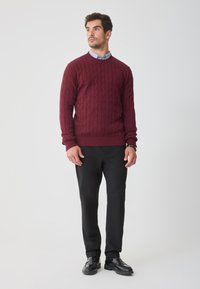 Bordowy sweter w splot kablowy z okrągłym dekoltem, noszony na niebieskiej koszuli z kołnierzykiem, w połączeniu z czarnymi spodniami i czarnymi butami.