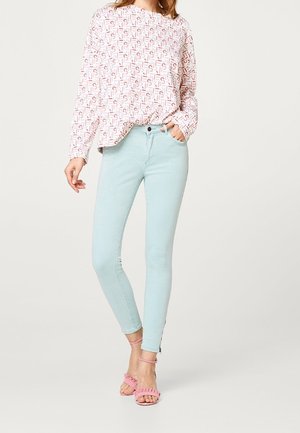 Nainen, jolla on vaaleansiniset kapeat farkut, valkoinen pitkähihainen paita punaisella kuviolla ja vaaleanpunaiset röyhelöiset avoimet korkokengät.