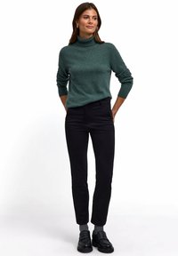 Dunkelgrüner Rollkragenpullover mit gerippten Bündchen, kombiniert mit schwarzen Slim-Fit-Hosen und schwarzen Slippern, auf einem Modell vor einem weißen Hintergrund.