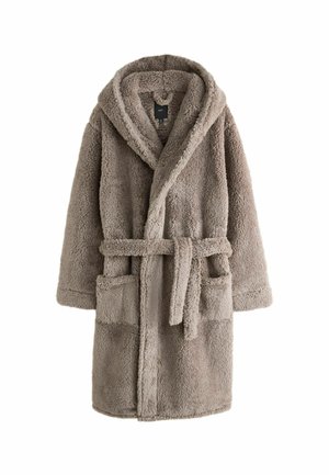Homme barbu portant un manteau en polaire épaisse beige texturée avec un grand col, sur un fond gris uni.