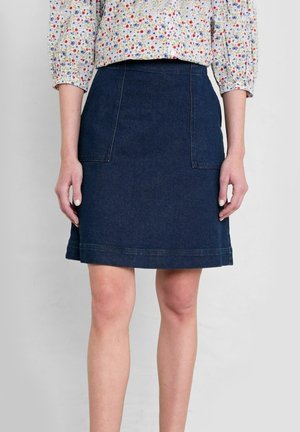 Femme portant une jupe trapèze en denim bleu foncé avec des poches plaquées à l'avant et un chemisier à manches longues fleuri, debout devant un fond uni.