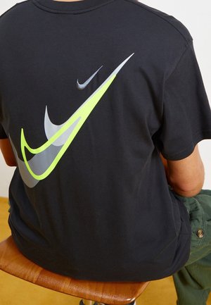 Personne portant un t-shirt noir avec de grands logos Nike swoosh gris, jaune et blanc superposés dans le dos, assise sur un tabouret en bois.