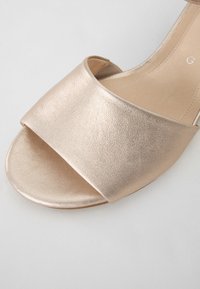 Avoin varvas sandaali metallinhohtoisessa rose gold -tekonahassa. Siinä on leveä, kaareva nauha jalan yli ja sileä, hieman teksturoitu pinta.