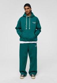Teal katoenen hoodie met kangoeroezak, witte trekkoorden en logo. Gecombineerd met bijpassende losse broek en zwarte sneakers.
