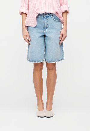 Person som har på seg lyseblå denim shorts til knærne, rosa stripete skjorte med knapper og hvite hælsko, står mot en hvit bakgrunn.