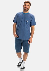 Modrá pruhovaná tričko s krátkým rukávem a kulatým výstřihem, s malou nášivkou loga, v kombinaci s džínovými šortkami a černými teniskami.