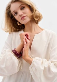 Helle Cremet Bluse mit gepufften Ärmeln und V-Ausschnitt. Ein burgunderfarbener Armreif am Handgelenk, minimale Textur und keine sichtbaren Verzierungen.