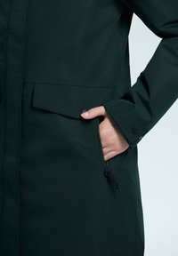 Chaqueta de lluvia verde con un bolsillo lateral con cremallera, cierre de botones a presión y una textura suave. De longitud media con un diseño de corte recto.