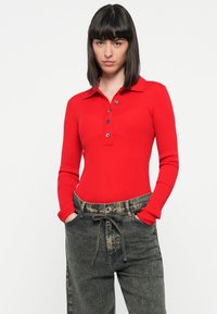 Polo à manches longues côtelé rouge avec un col, doté d'une patte de boutonnage. Assorti à un pantalon en denim noir usé à taille haute.
