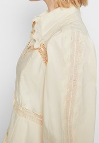 Woman wearing cream blouse with scalloped lace trim on collar, shoulder, and sleeve, shown from side at neck and upper torso.