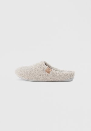Beige pantoffel met een gestructureerde, fuzzy buitenkant, instapontwerp en een klein leren accentlabel aan de zijkant. Zachte voering en rubberen zool.