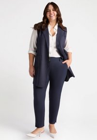 Marineblauwe op maat gemaakte vest over een witte blouse, gecombineerd met smal gesneden marineblauwe broek en witte hakken; gladde stof en gestructureerd ontwerp.