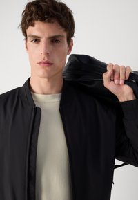 Black bomber jacket worn over a light beige textured shirt. Smooth fabric with ribbed cuffs; model holding a dark bag on shoulder.