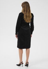 Svart manchesterklänning med långa ärmar, figursydd siluett och knälång fåll. Bärs med svarta slingback-klackar.