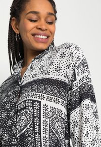 Camisa com padrão preto e branco, apresentando designs florais e geométricos, com gola e detalhes em botão. Tecido macio.