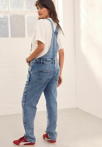 Overoles de mezclilla con un corte relajado, color azul claro, tirantes ajustables y bolsillos frontales, combinados con una camiseta blanca y zapatillas rojas.