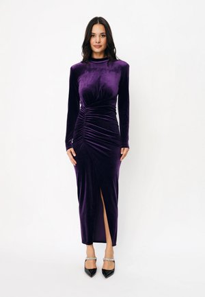 Une femme présente une robe en velours violet à manches longues, avec une taille froncée et une fente à l’avant, portée avec des talons noirs sur fond blanc.
