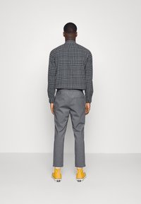 Hombre de espaldas vistiendo una camisa de cuadros en negro y gris, pantalones grises, calcetines blancos y zapatillas altas de color amarillo brillante.