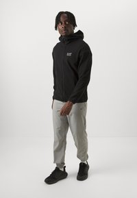 Svart zip-up hettegenser med høy krage, med hvit EA7-logo. Brukt med lysegrå bukser og svarte joggesko. Glatt stofftekstur.