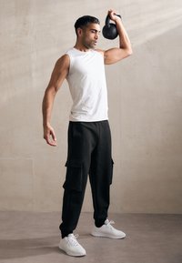 Mann hebt mit dem rechten Arm eine schwarze Kettlebell über den Kopf, trägt ein weißes ärmelloses Shirt, schwarze Cargohose und weiße Sneakers, steht auf grauem Boden.