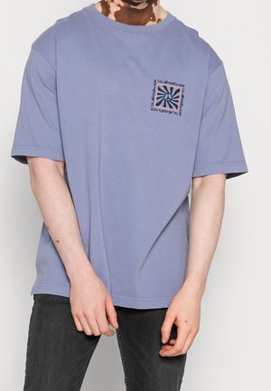 Mężczyzna ubrany w luźną lawendową koszulkę z krótkim rękawem, z małym czarno-białym kwadratowym nadrukiem i napisem Quiksilver na lewej piersi.