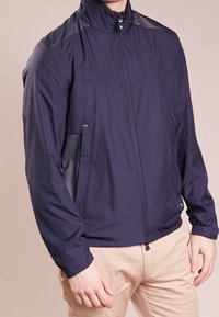 Mörkblå lättviktsjacka med mesh-detaljer, dragkedjestängning, sidofickor och elastiska muddars, designad för komfort och andningsförmåga.