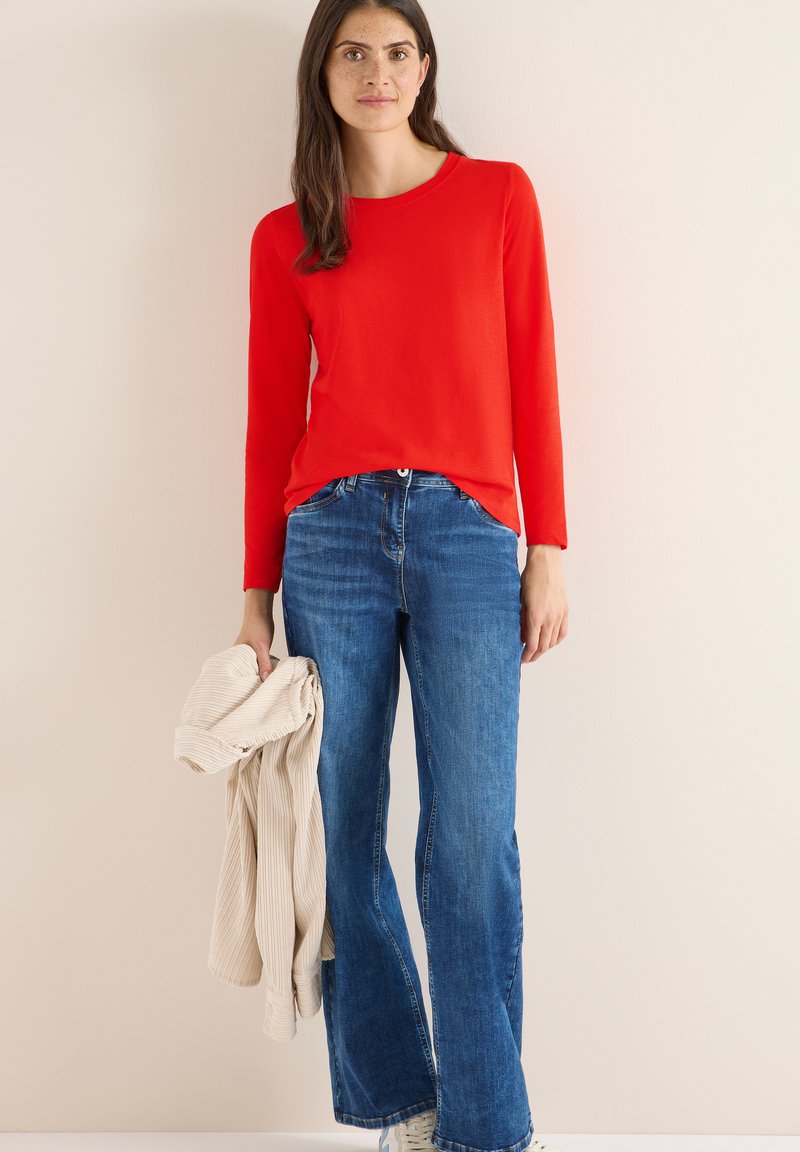 Rotes langärmliges Oberteil kombiniert mit blauen Schlagjeans. Das Model hält ein hellbeiges strukturiertes Hemd. Minimalistischer Hintergrund, der die Details der Kleidung zeigt.