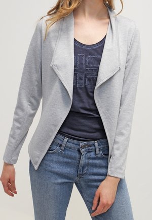Chaqueta gris claro de mangas largas con un diseño drapeado en la parte delantera y una textura sutil, combinada con una blusa azul marino de cuello redondo y pantalones vaqueros azules.