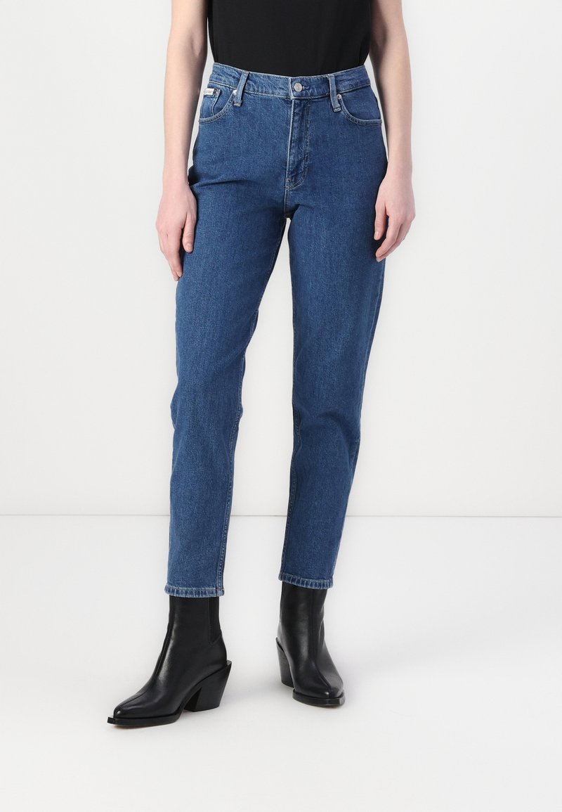 Jean en denim bleu avec une taille haute, un design à jambe droite et des détails de poches avant cousus. Associé avec des bottines à talons noires.