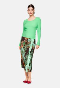 Grüner, strukturierter Langarmoberteil kombiniert mit einem braunen Rock im Blumen- und Karomuster, ergänzt durch rote flache Schuhe.