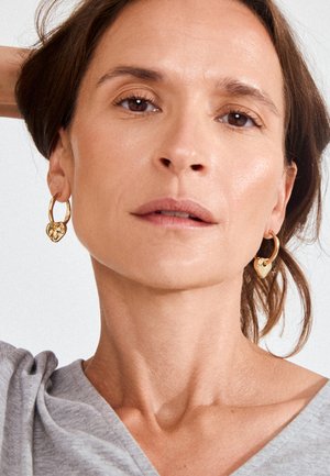 Close-up of a woman with light brown hair wearing gold hoop earrings with heart-shaped charms, a gray top, and a neutral expression.