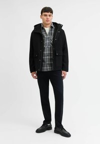 Chaqueta negra con capucha, camisa de cuadros gris, pantalones negros y zapatos negros. La chaqueta tiene bolsillos frontales y cierre de cremallera.
