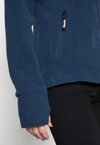 Navy blue fleece jacket with a high collar, front zip pocket, and long, wide sleeves, paired with black pants. Soft, textured fabric.