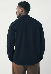 Camisa de un azul marino oscuro con cuello estructurado y mangas largas. Presenta una parte trasera recta con detalles de costura sutiles. Textura suave.