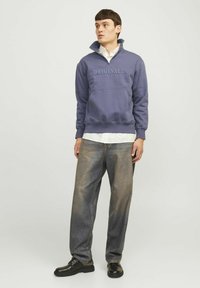 Felpa viola con colletto alto, con la scritta "ORIGINALS", abbinata a jeans baggy blu e scarpe nere.