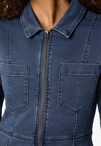 Denim overall i mørkeblå med glidelås foran, spredt krage og to brystlommer; har stikking detaljer og en tilpasset design.