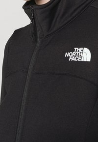 Czarna polarowa kurtka z wysokim kołnierzem, z zamkiem błyskawicznym oraz białym logo The North Face po lewej stronie klatki piersiowej. Miękka tekstura.