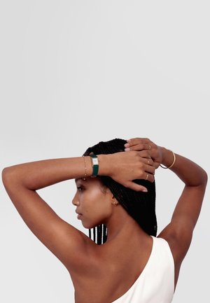 Žena s copánky, oblečená v bílém topu, pózující s rukama zvednutýma za hlavou, nosící zelené hodinky a zlaté šperky.