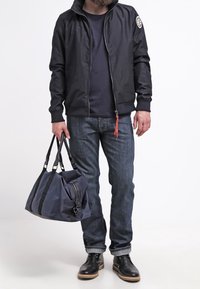 Marinblå bomberjacka med dragkedja i fram, kombinerad med mörka jeans och svarta läderstövlar. Bär en stor blå väska med svarta remmar och dragkedja.