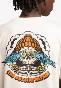Camiseta color crema con un gráfico colorido de una calavera que lleva un gorro naranja, flanqueada por pájaros azules. El texto dice "EL EXTERIOR DENTRO".