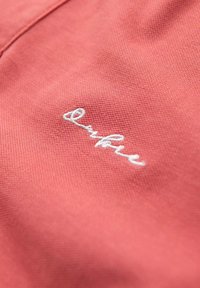 Koralli kangas, jossa on tekstuuripinta, sisältää valkoisen brodeeratun logon. Lähikuva korostaa ompeluyksityiskohtia ja kirkasta väriä.